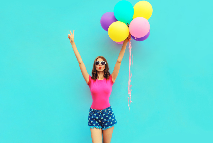 Beautiful young woman raising her hands up with bunch of balloons having fun wearing a shorts and pink t-shirt on blue wall background